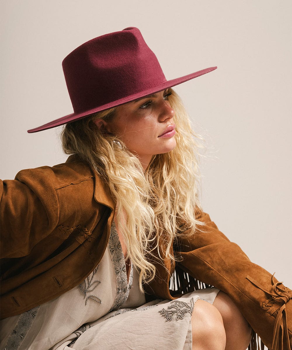 Woman wearing a merlot fedora hat and a dress with a brown coat, facing sideways against a plain background #color_merlot