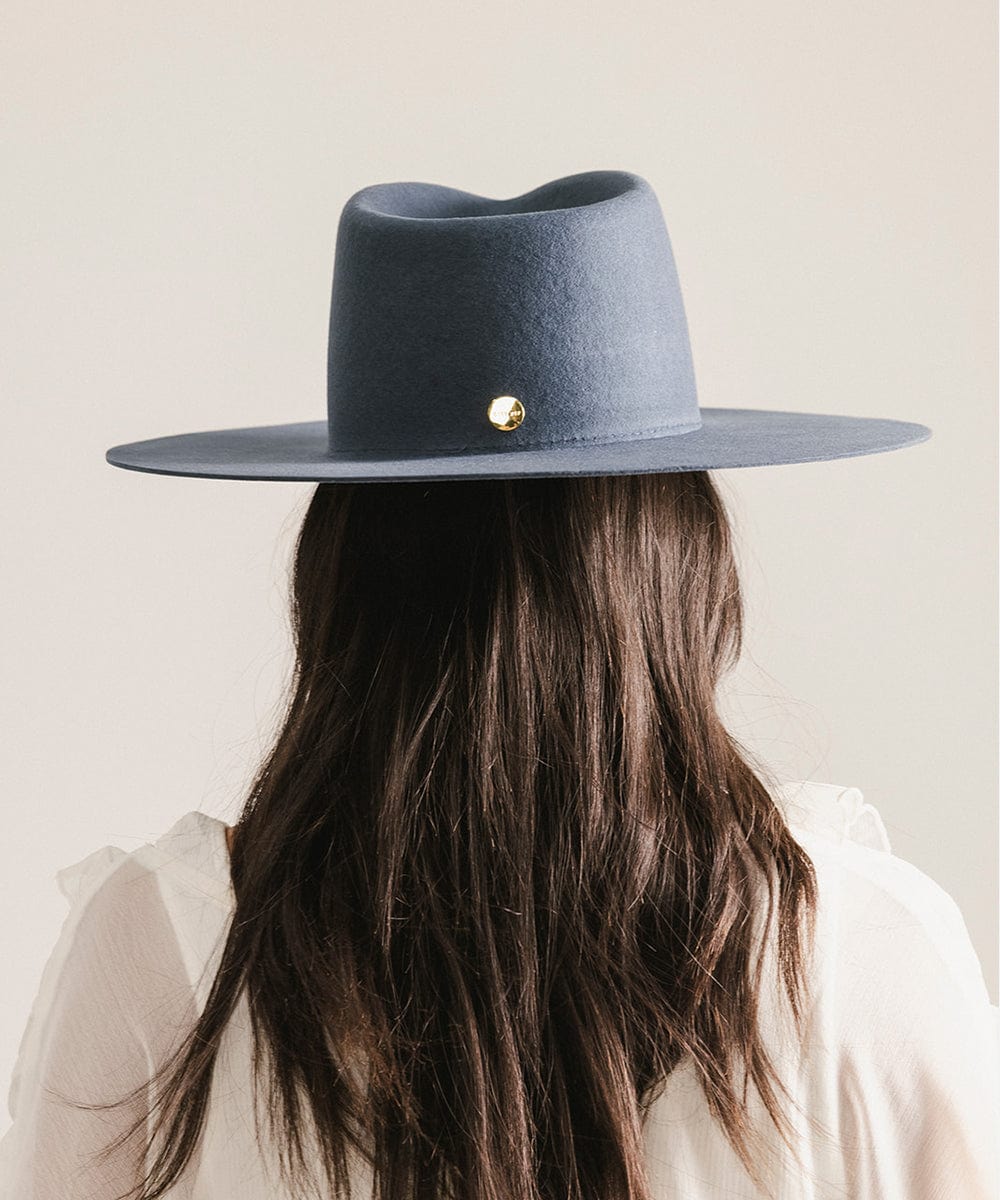 Woman wearing a vintage blue colored fedora hat and a white top, facing behind against a plain background #color_vintage blue