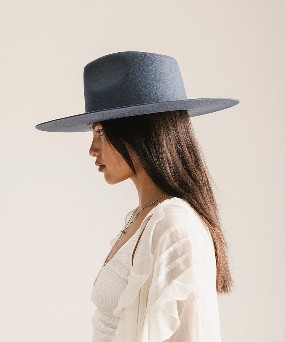 Woman wearing a vintage blue colored fedora hat and a white top, facing sideways against a plain background #color_vintage blue