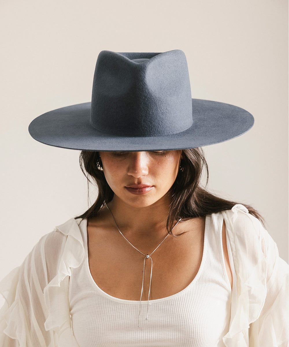 Woman wearing a vintage blue colored fedora hat and a white top, looking down against a plain background #color_vintage blue
