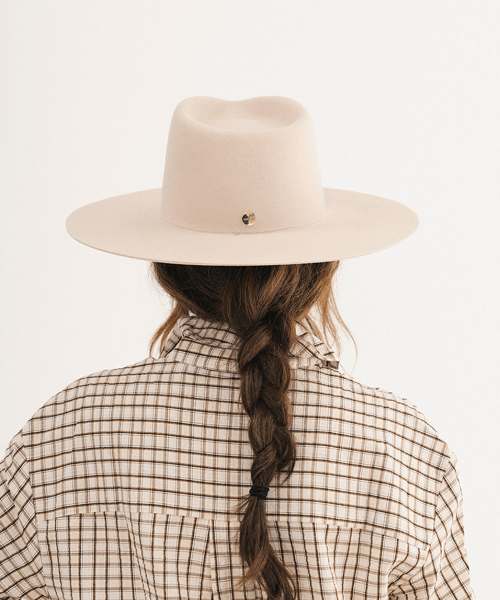 A person viewed from behind wearing a wide-brim fedora hat in a light blush beige color, paired with a brown and beige checkered shirt. The hat features a small gold heart-shaped pin on the back of the crown #color_cream