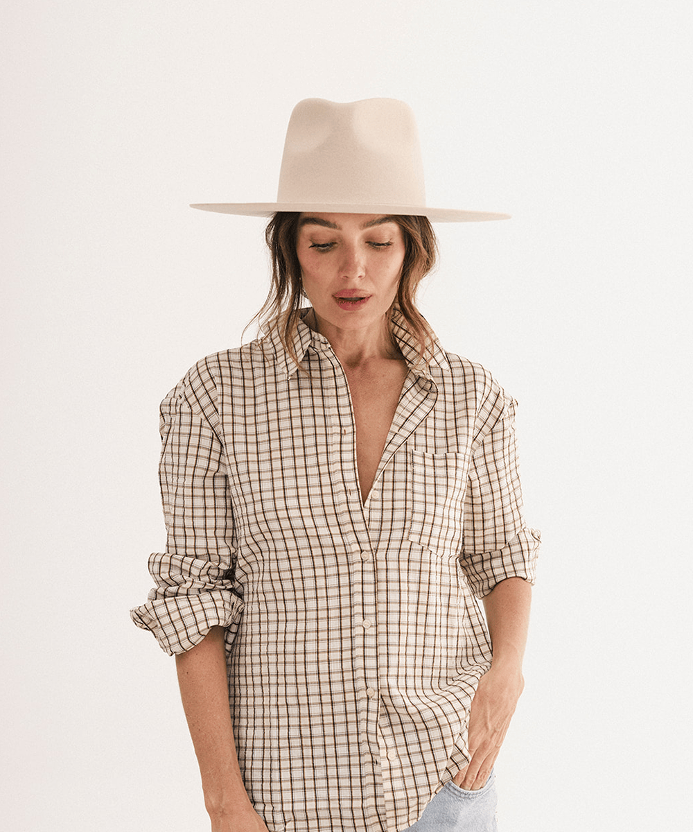 A woman wearing a beige wide-brim fedora hat and a brown and beige checkered button-down shirt, paired with light blue jeans, photographed against a clean white studio background #color_cream
