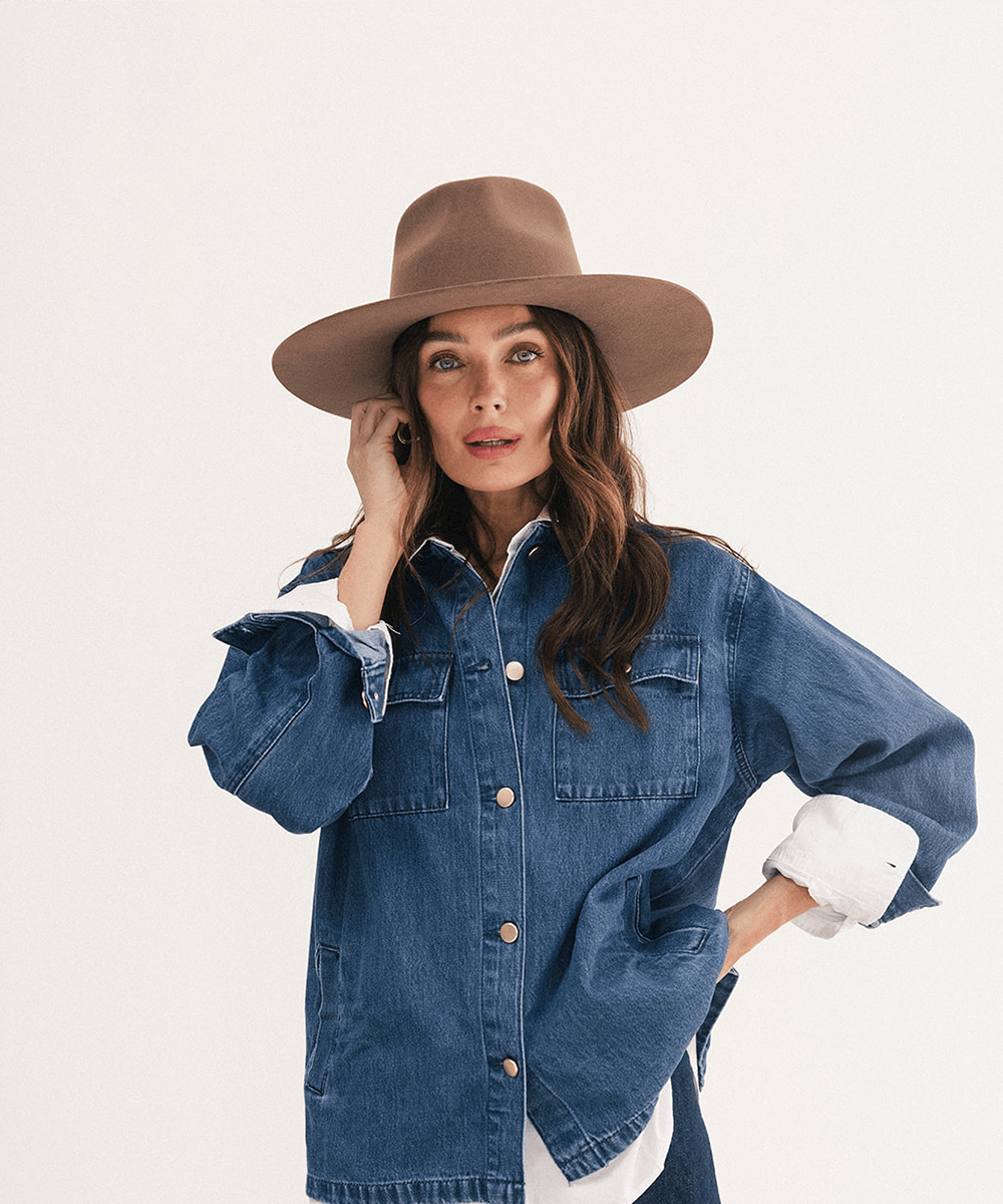 A woman wearing a brown wide-brim fedora hat and an oversized blue denim shirt jacket with rolled-up sleeves, posed against a clean off-white studio background #color_chocolate