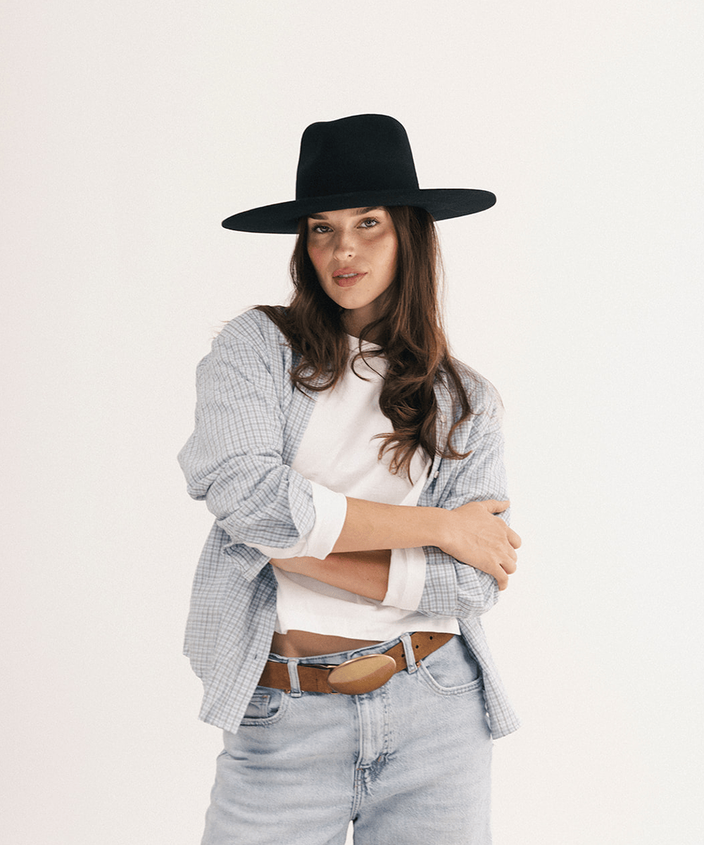 A woman modeling a black wide-brim fedora hat, styled with a light blue and white checkered shirt, a white t-shirt, light-wash jeans, and a brown leather belt against a plain white studio background #color_black