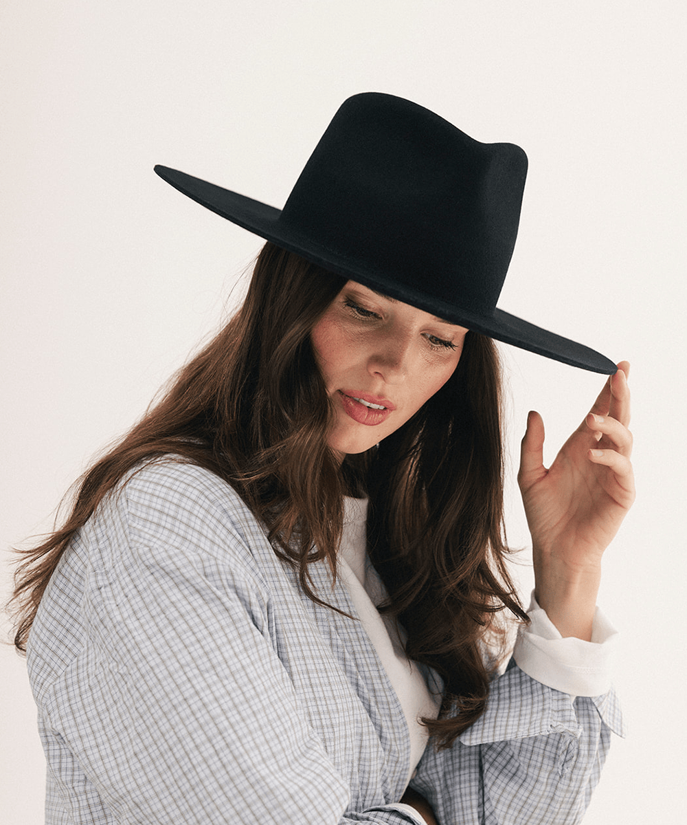 A woman wearing a black wide-brim fedora hat, styled with a light blue and white checkered shirt against a plain off-white background #color_black