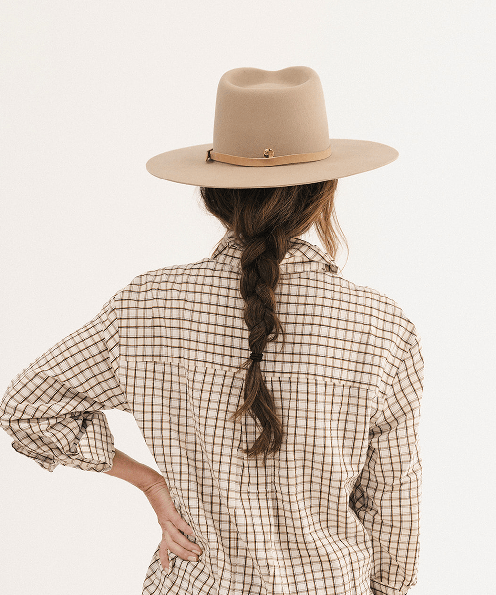A woman shown from behind wearing a beige wide-brim fedora hat with a tan band and a beige-and-brown plaid button-up shirt, against a plain white studio background #color_tan