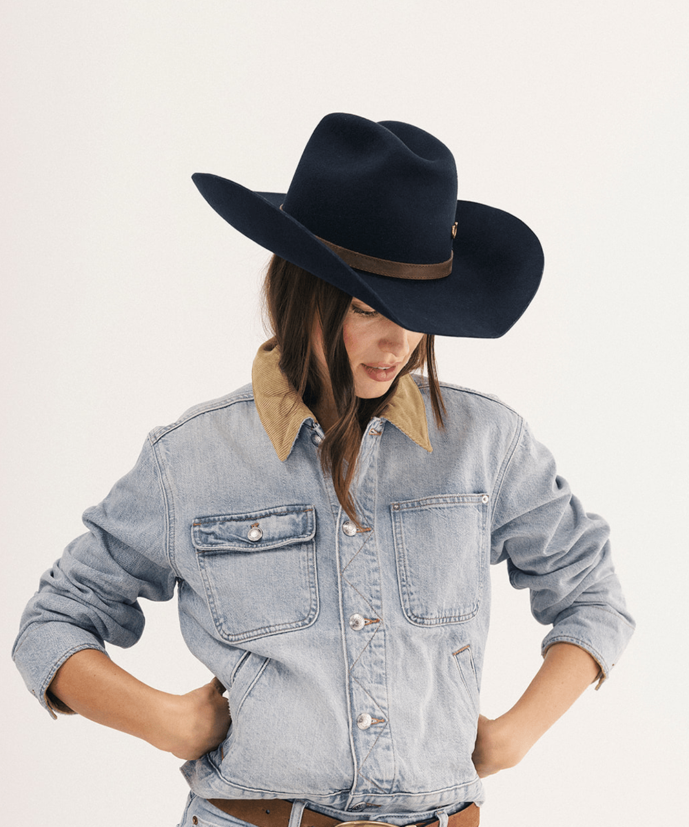 A woman wearing a dark navy blue felt cowboy hat with a brown band against a plain light background #color_navy