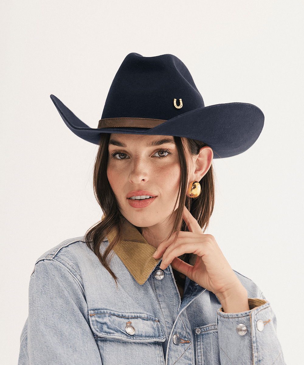 A woman wearing a dark navy blue felt cowboy hat with a brown band and a small gold horseshoe emblem, paired with a light blue denim jacket and gold hoop earrings, against a plain white background #color_navy