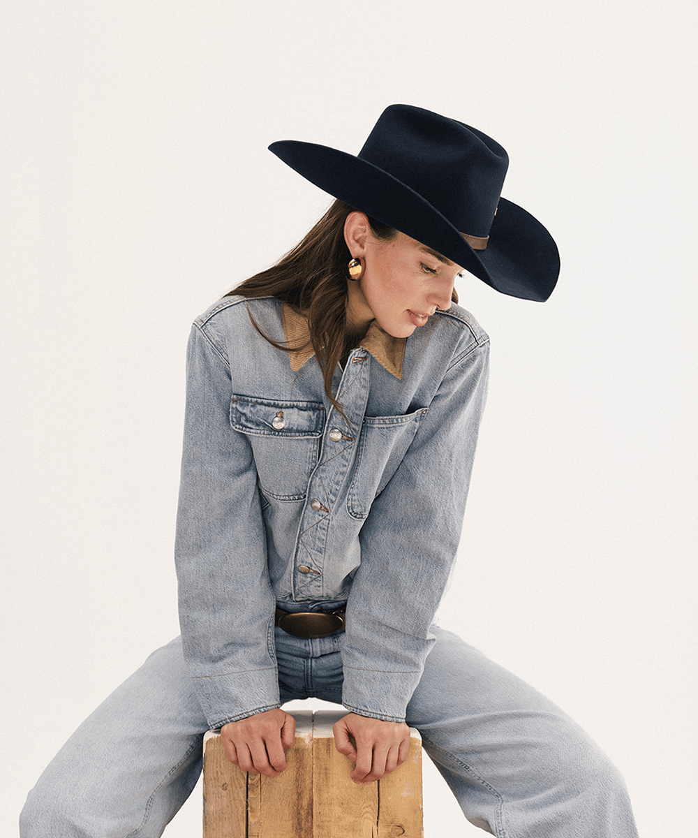 A woman wearing a black cowboy hat, a light blue denim shirt, and matching jeans sits on a wooden crate against a plain white studio background #color_navy