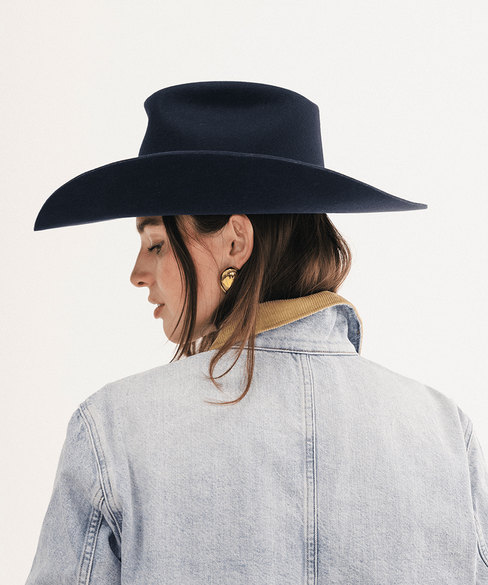 A woman shown from the back wearing a dark navy wide-brim fedora hat, a light blue denim jacket with a tan collar, and a gold heart-shaped earring, against a plain white background #color_navy