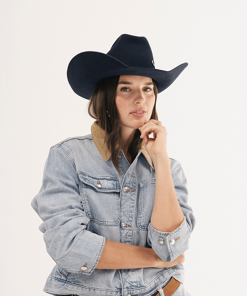 A woman wearing a black cowboy hat and a light-wash denim jacket with a tan collar, posing against a plain white studio background #color_navy