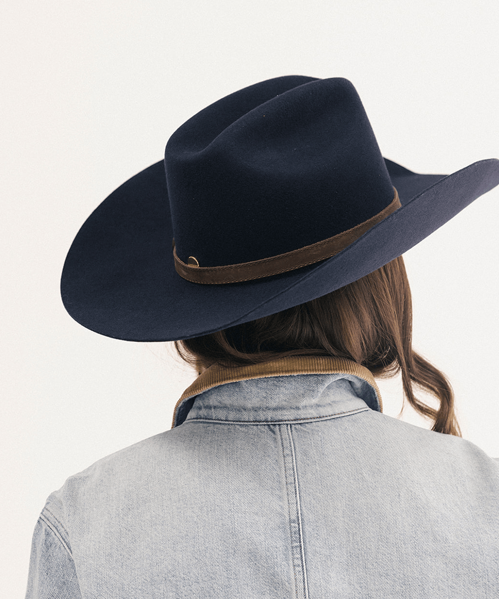 A person shown from behind wearing a dark navy blue felt cowboy hat with a brown band, a light denim jacket, and brown hair, against a plain off-white background #color_navy