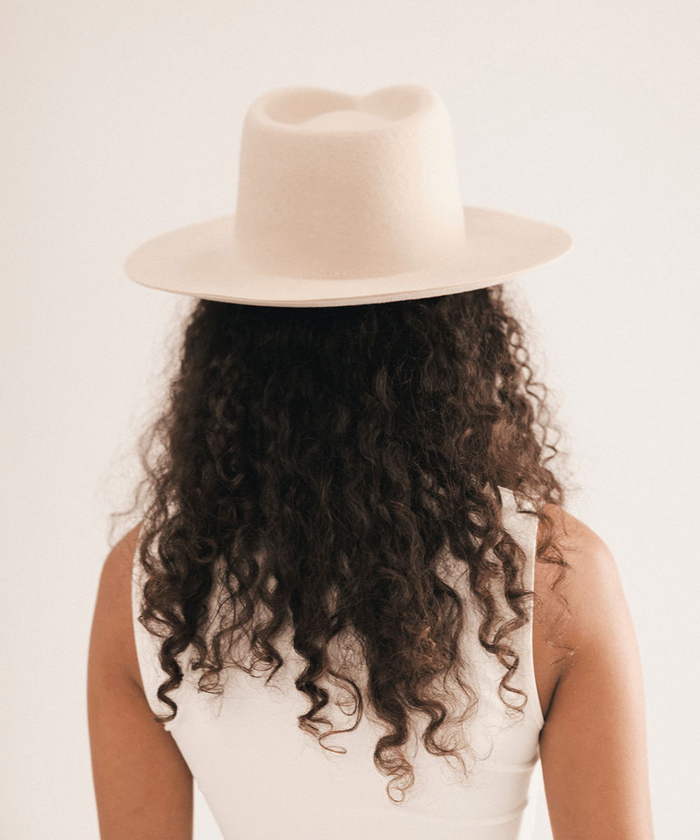 A person shown from the back wearing a light cream-colored felt fedora hat and a white sleeveless top, with long dark curly hair visible #color_cream