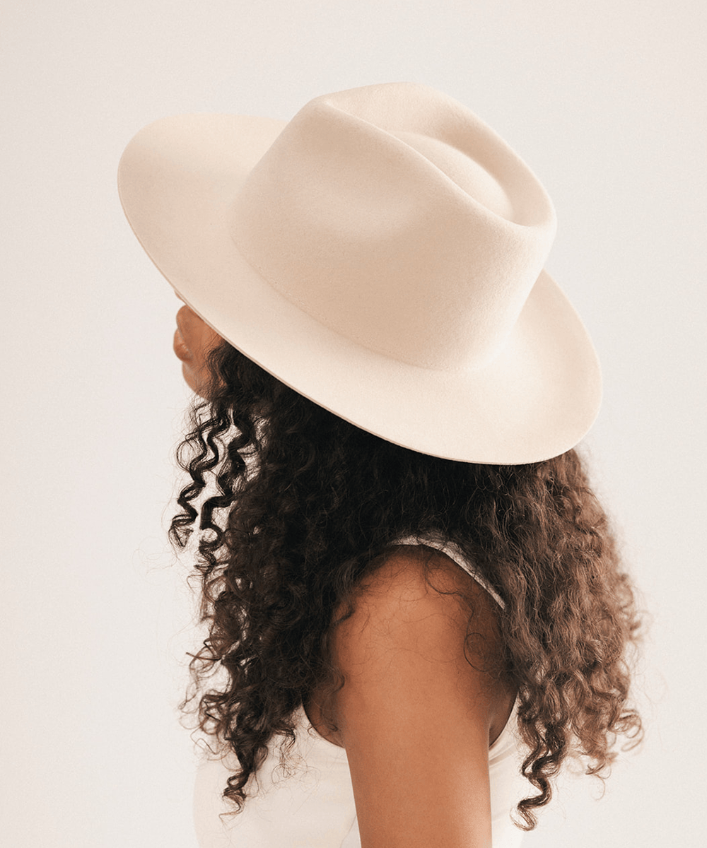 A woman shown from the side wearing a wide-brim fedora hat in a solid cream color, with her dark curly hair visible #color_cream