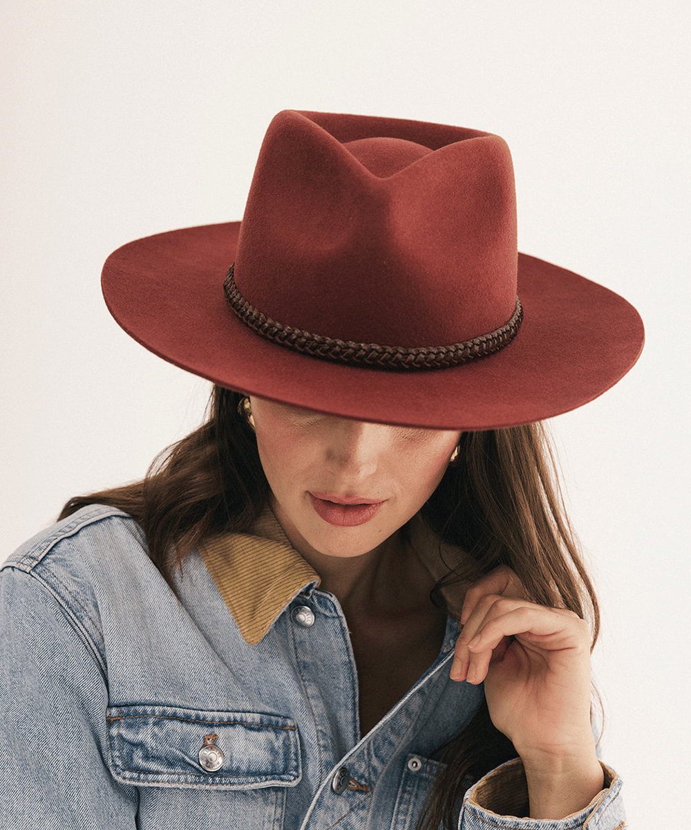 A woman wearing a wide-brimmed rust-colored felt hat with a braided dark band and a light blue denim jacket, photographed against a clean off-white background #color_rusty red