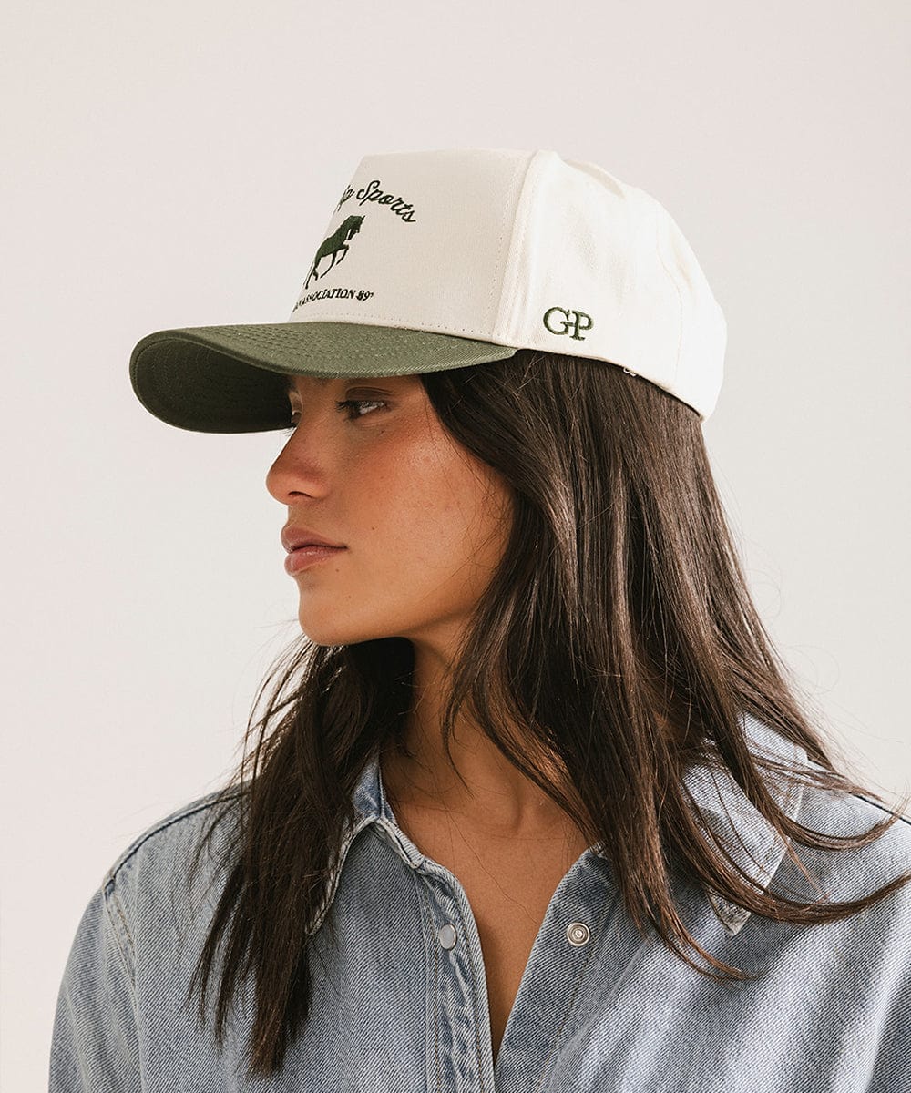 Woman wearing a cream and dark green Trucker hat with 'Gigi Pip Sports' and horse logo and a denim shirt, facing sideways against a plain background #color_cream - dark green