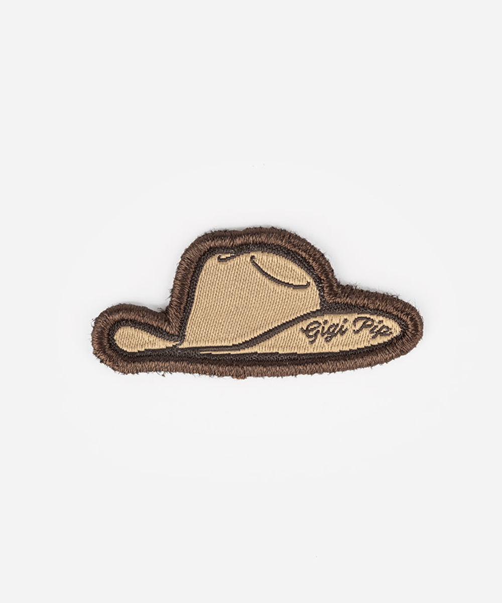 An embroidered patch shaped like a cowboy hat, featuring a tan center with a dark brown stitched border and script text reading 'Gigi Pipi' on the right side, set against a plain light gray background #color_tan