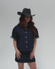 Woman wearing a dark grey cowboy hat with an upturned brim and a golden button and an attached leather band, and a dark blue denim shirt and a denim skirt, posing against a white background #color_dark grey