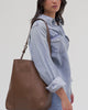 Woman wearing a blue denim shirt and blue jeans, carrying a chocolate colored Dia everyday leather tote bag, posing against a plain background #color_chocolate