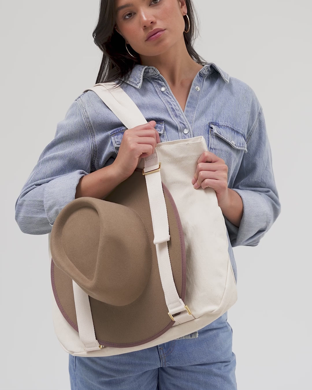 Woman wearing a denim shirt carrying a hat on a natural colored canvas tote bag against a plain background #color_natural