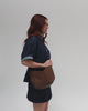 Woman wearing a denim shirt and a denim skirt, carrying a chocolate colored genuine leather slouchy bag against a plain background #color_chocolate