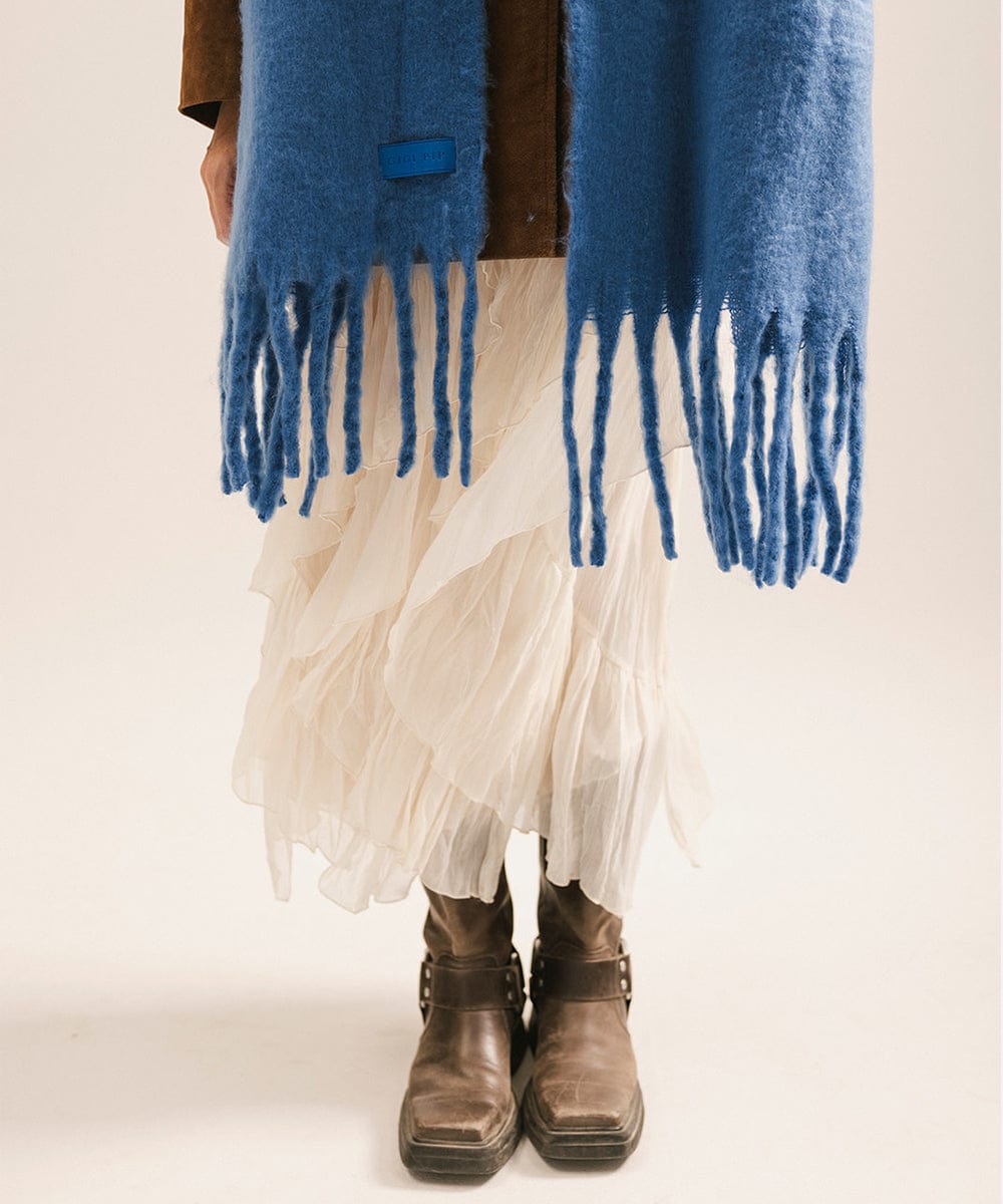 Person wearing a blue scarf with fringe, white pants, and brown boots on a plain background #color_alpine blue