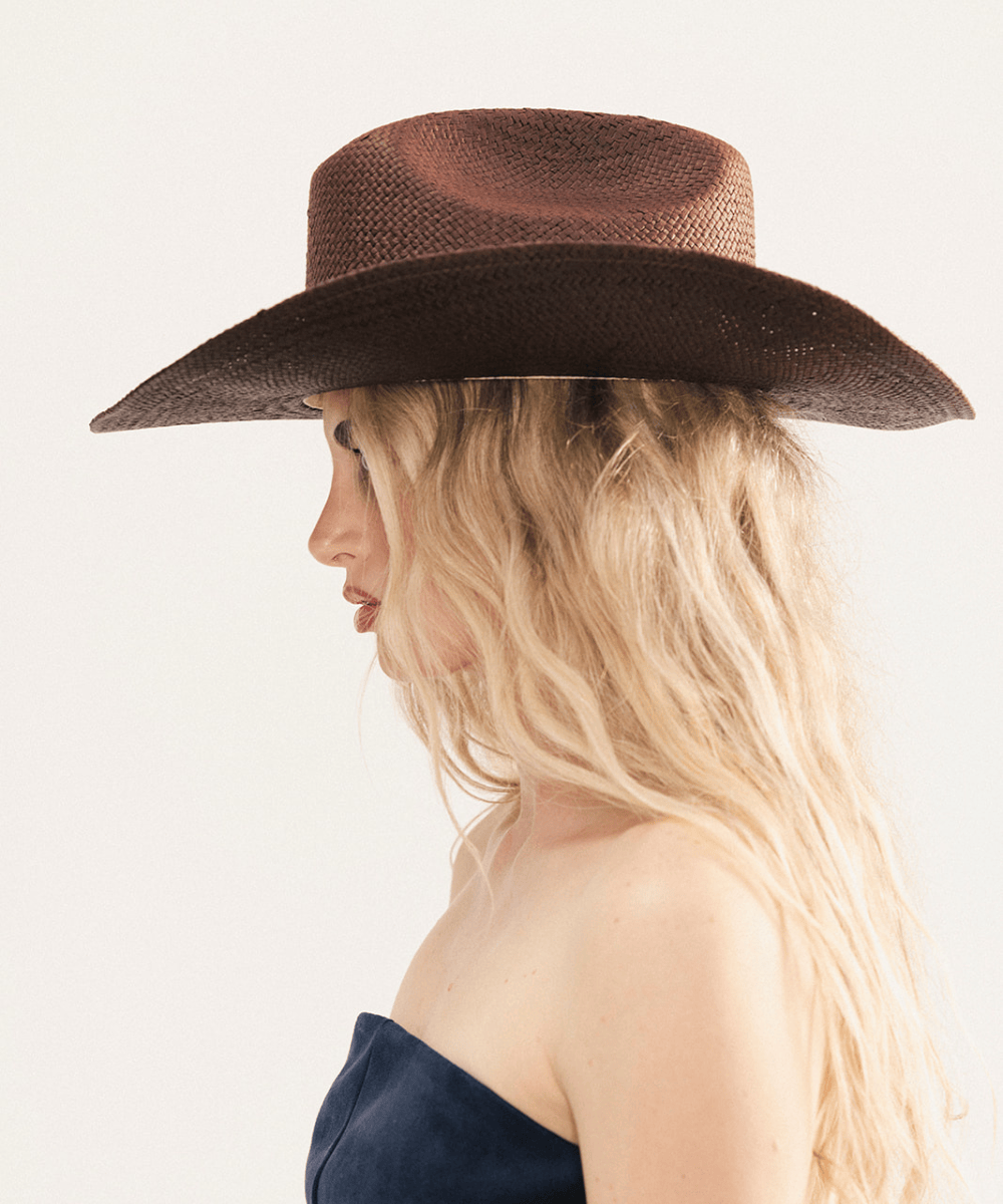 Woman wearing a dark brown straw cowboy hat and dark clothing against a plain background #color_dark brown