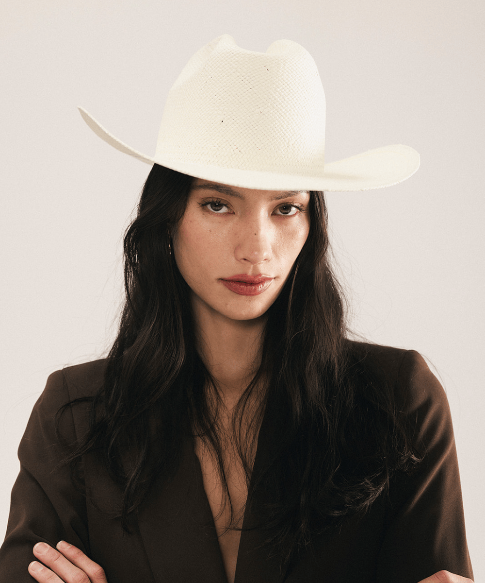 Woman wearing a white straw cowboy hat and dark clothing against a plain background #color_natural white