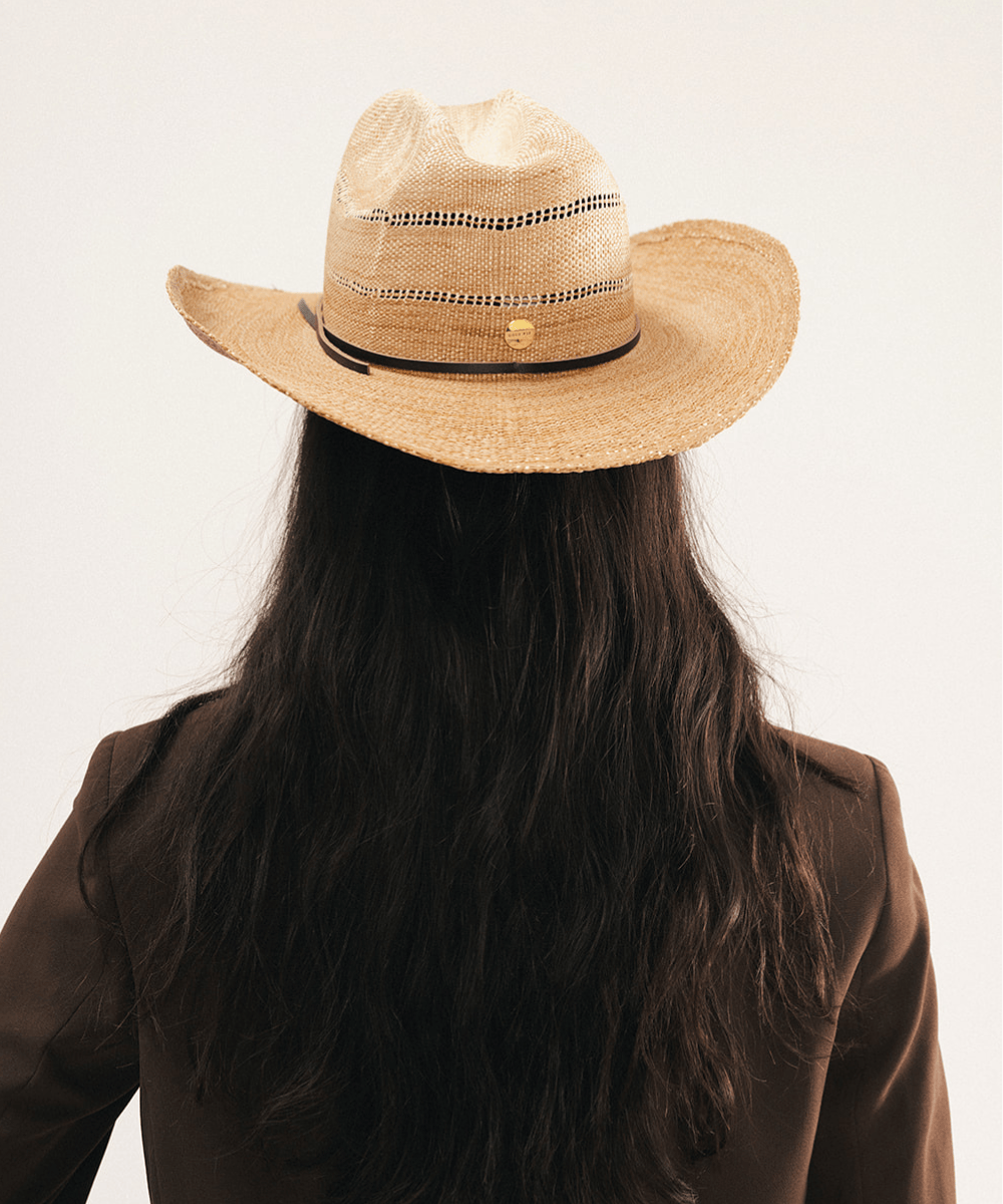 Woman wearing a washed tan straw cowboy hat and dark clothing against a plain background #color_washed tan