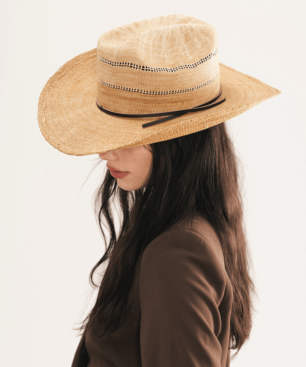 Woman wearing a washed tan straw cowboy hat and dark clothing against a plain background #color_washed tan