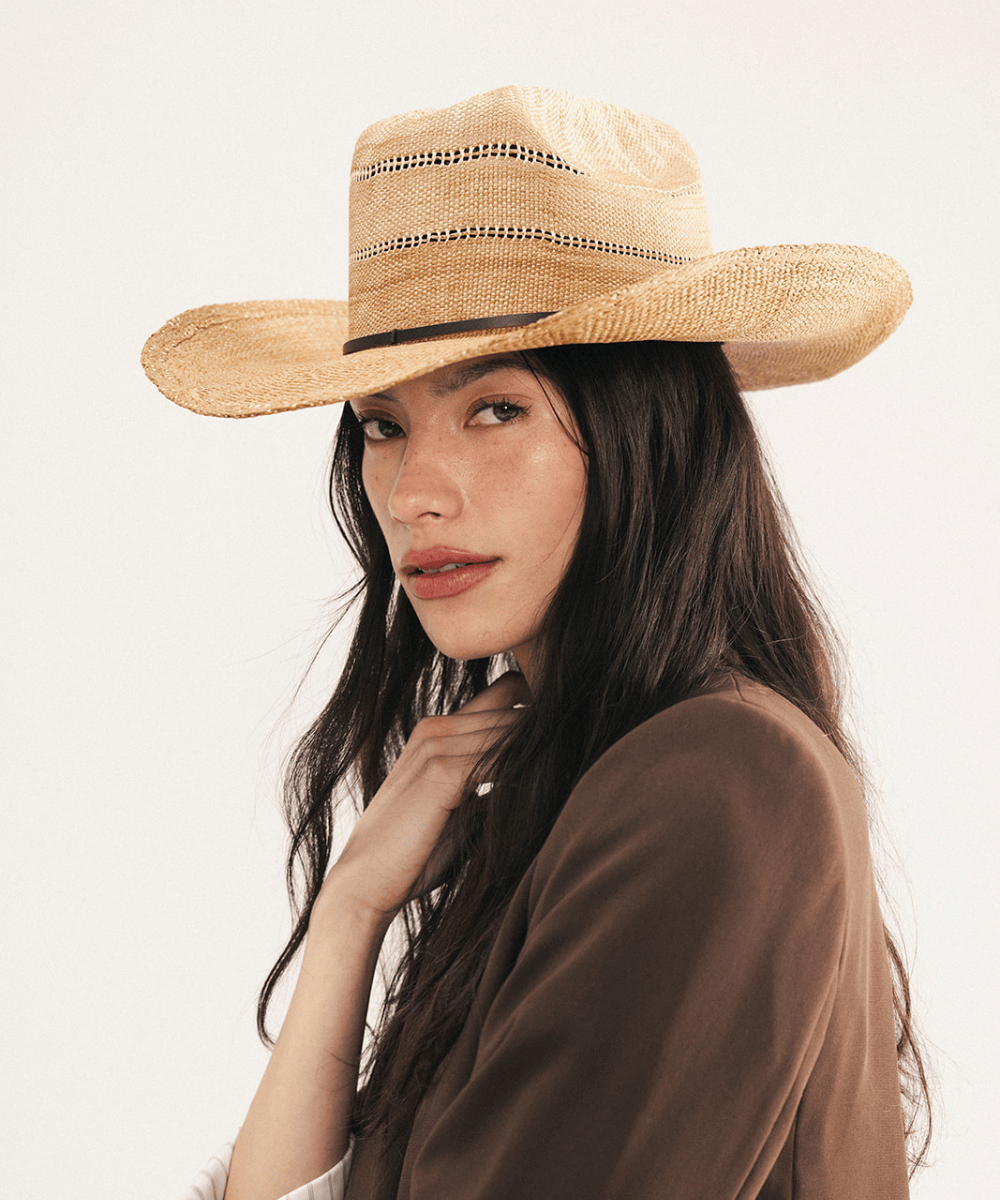 Woman wearing a washed tan straw cowboy hat and dark clothing against a plain background #color_washed tan