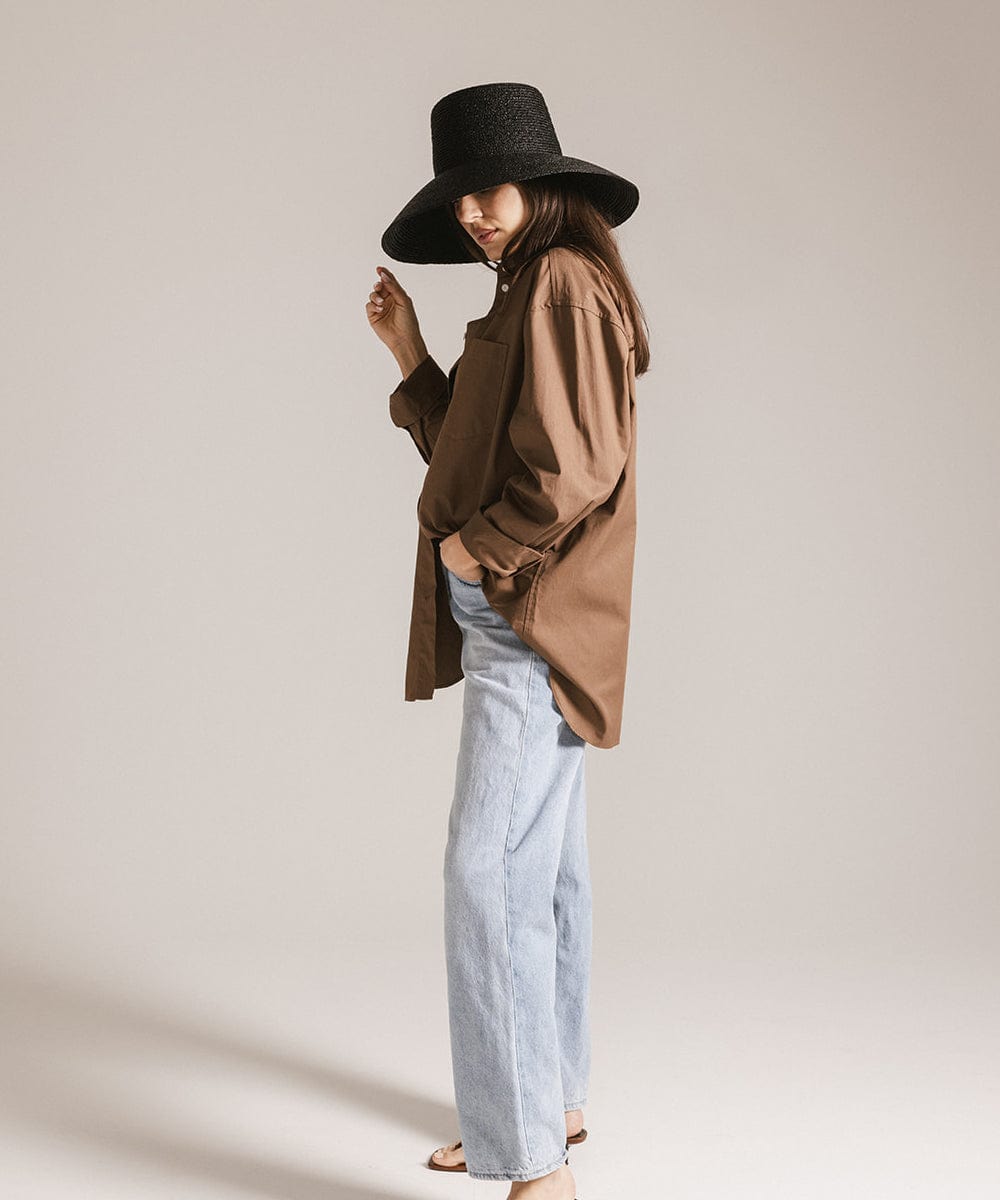 Woman wearing a black oversized raffia straw bucket hat, a brown shirt and light blue jeans against a plain background #color_black 