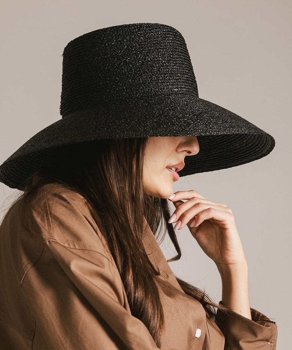 Woman wearing a black oversized raffia straw bucket hat and a brown shirt, facing sideways against a plain background #color_black 