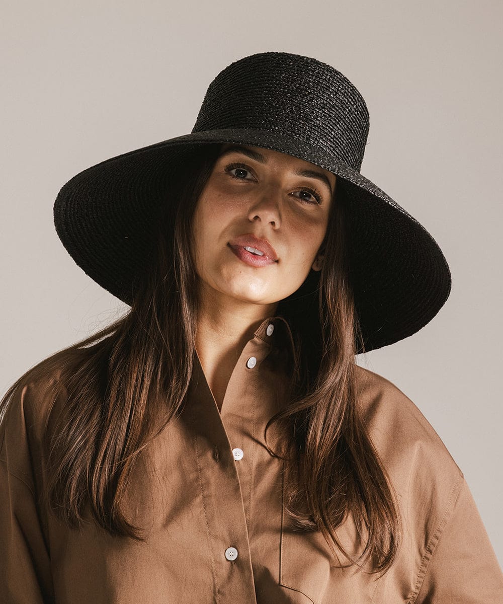 Woman with brown hair wearing a black oversized raffia straw bucket hat and a brown shirt against a plain background #color_black 