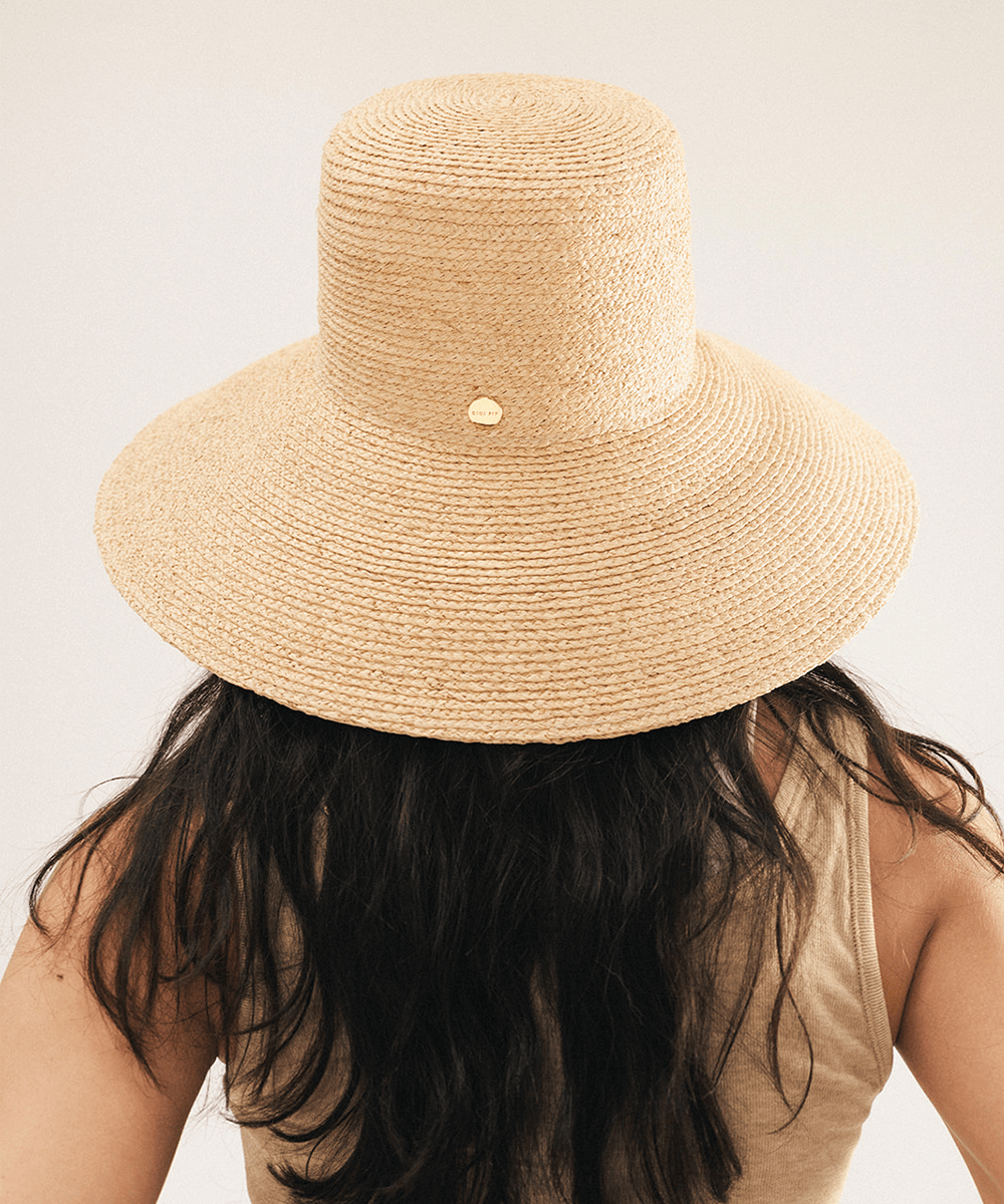Woman wearing a natural colored oversized raffia straw bucket hat with a wide brim and sunglasses on a plain background #color_natural