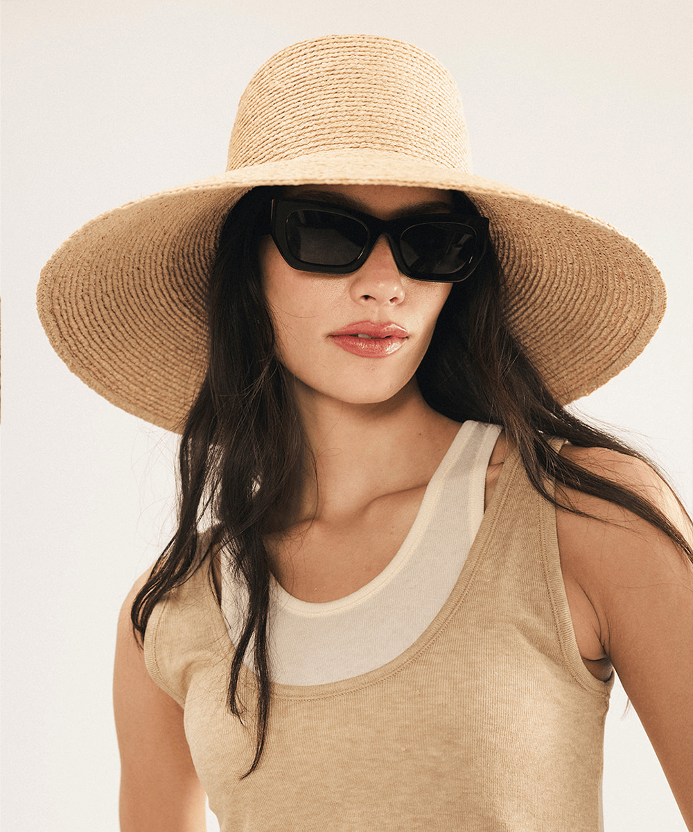 Woman wearing a natural colored oversized raffia straw bucket hat with a wide brim and sunglasses on a plain background #color_natural