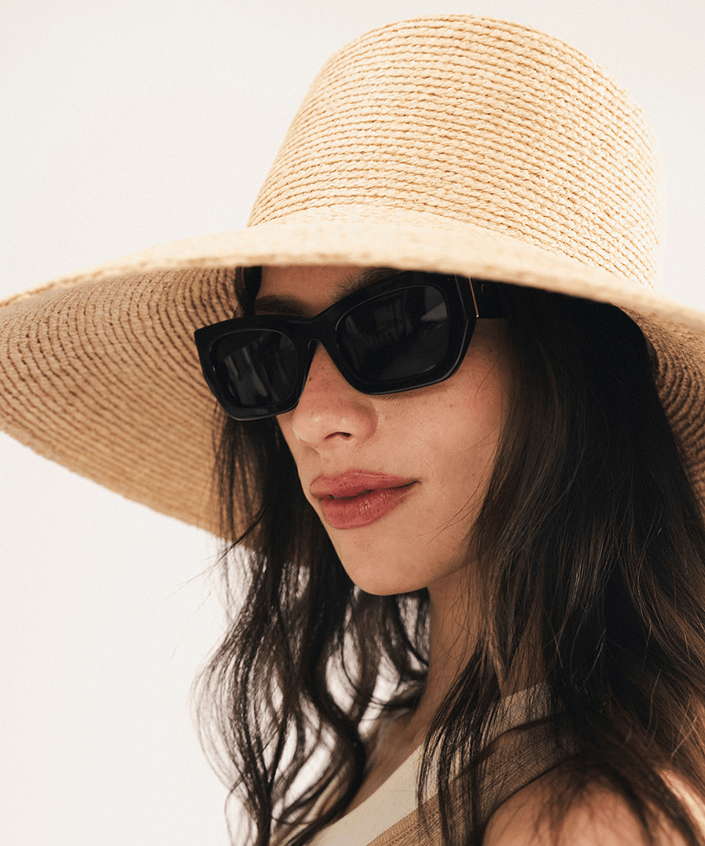 Woman wearing a natural colored oversized raffia straw bucket hat with a wide brim and sunglasses on a plain background #color_natural