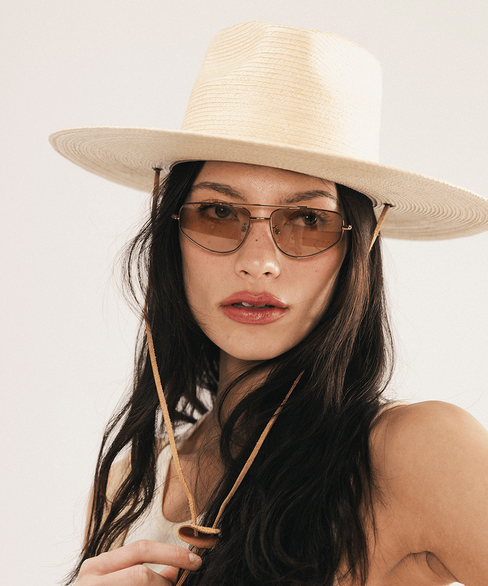 Woman wearing an ivory colored palm straw sun hat with a teardrop fedora crown and a wide flat brim, featuring a brown genuine leather chinstrap on a white background #color_ivory