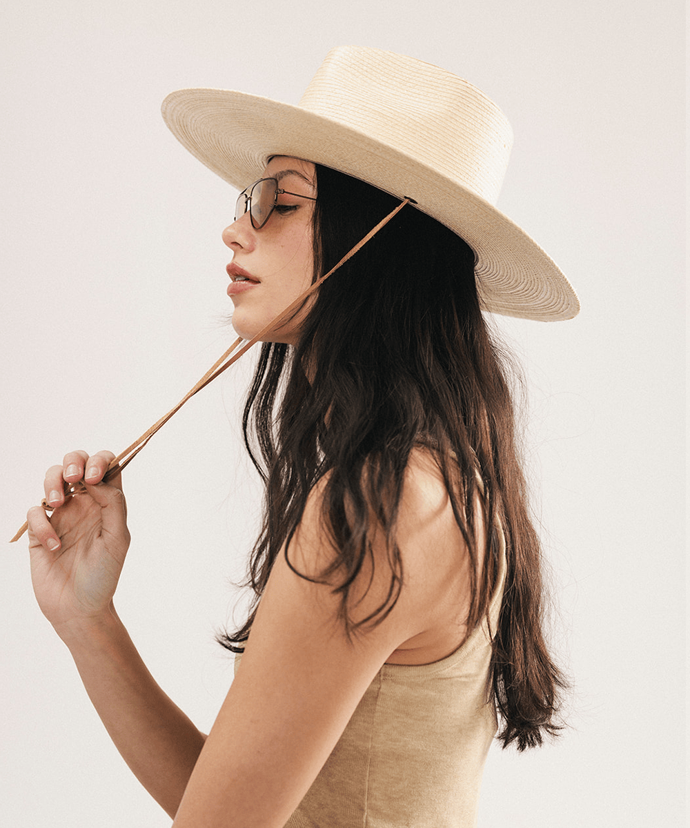 Woman wearing an ivory colored palm straw sun hat with a teardrop fedora crown and a wide flat brim, featuring a brown genuine leather chinstrap on a white background #color_ivory
