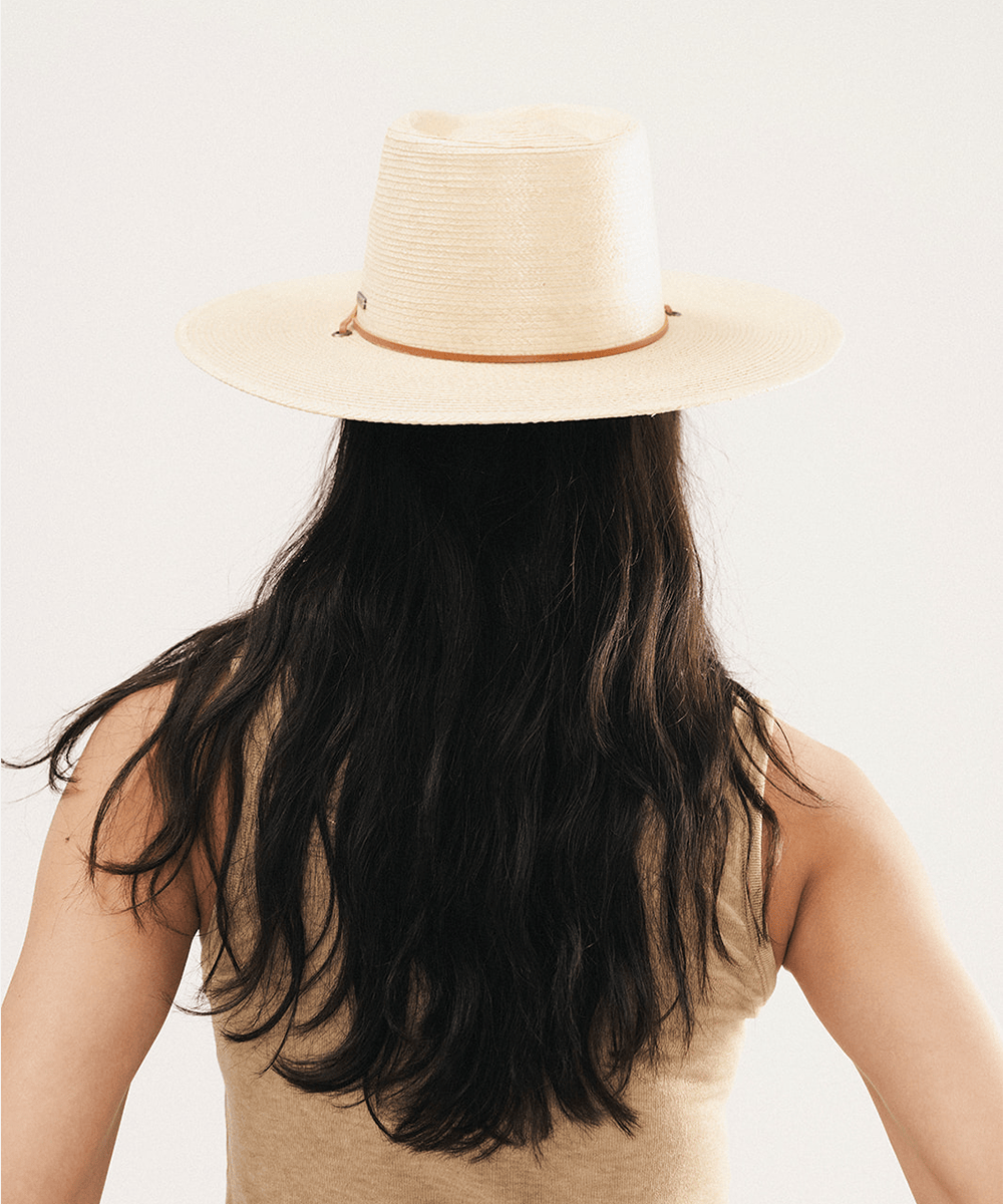 Woman wearing an ivory colored palm straw sun hat with a teardrop fedora crown and a wide flat brim, featuring a brown genuine leather chinstrap on a white background #color_ivory
