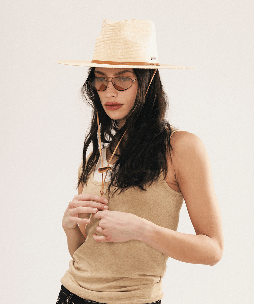Woman wearing an ivory colored palm straw sun hat with a teardrop fedora crown and a wide flat brim, featuring a brown genuine leather chinstrap on a white background #color_ivory