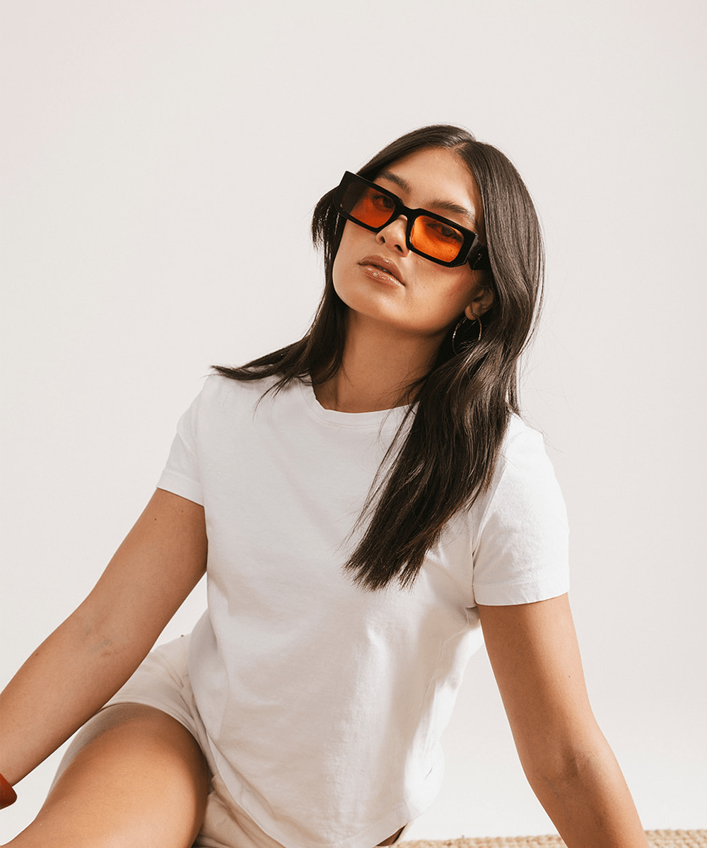 Woman wearing sunglasses with black frame and orange lenses and a white t-shirt against a plain background #color_black