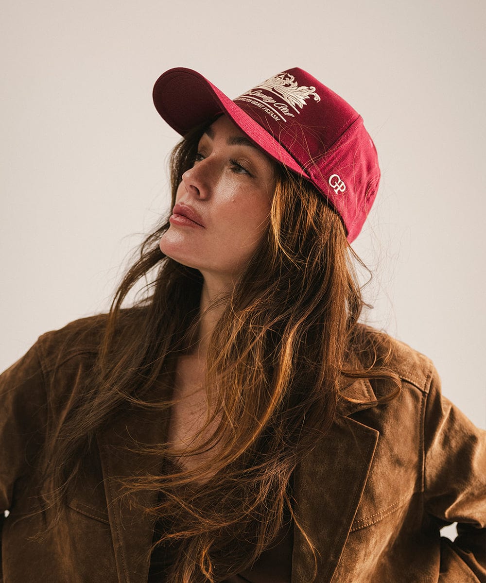 Woman wearing a burgundy vintage trucker cap with a white logo and a brown coat, posing against a plain background  #color_burgundy