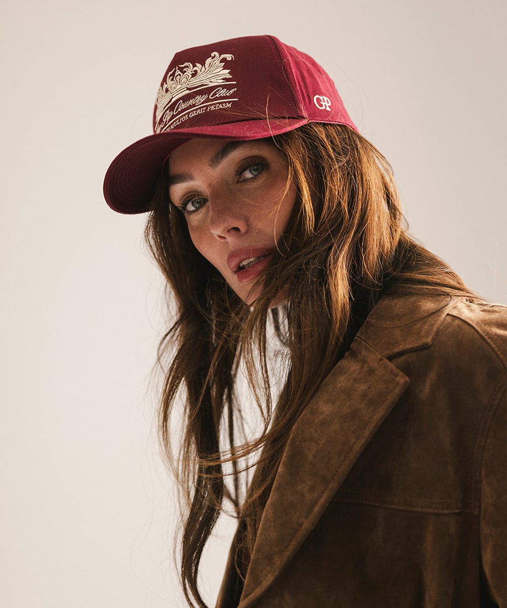 Woman wearing a burgundy vintage trucker cap with a white logo and a brown coat against a plain background  #color_burgundy
