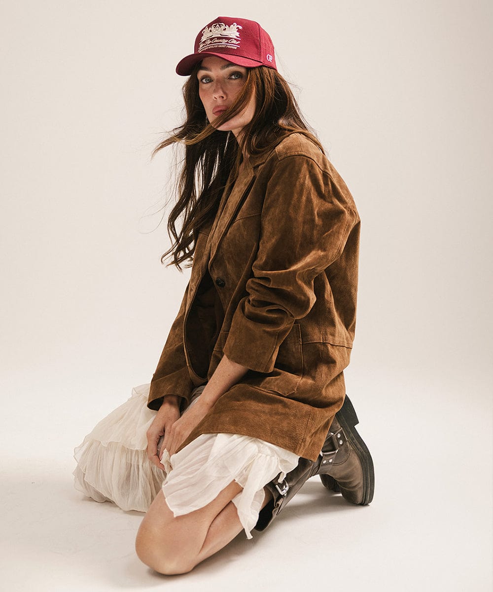 Woman wearing a burgundy vintage trucker cap with a white logo, a brown coat and a white skirt, sitting against a plain background  #color_burgundy
