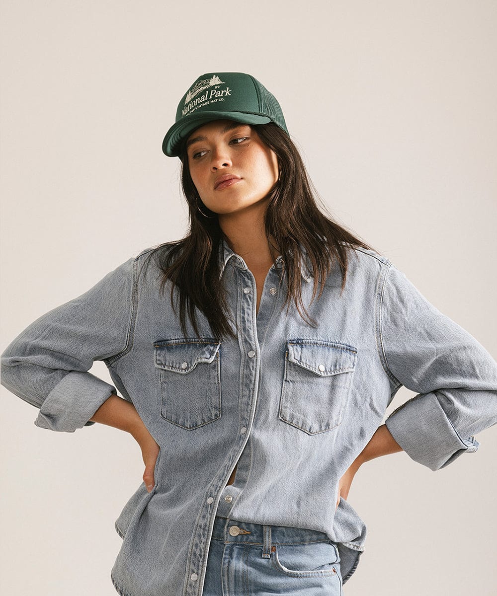 Woman wearing a forest green colored trucker hat with 'National Park' logo and a denim shirt, posing  against a plain background  #color_forest green