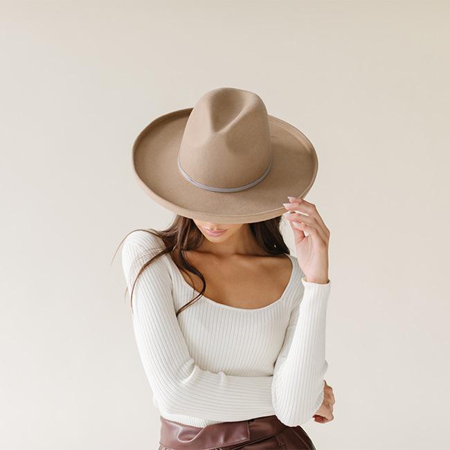 Woman wearing a tan pencil brim fedora hat with a hat band and a white top, looking down against a plain background #color_tan