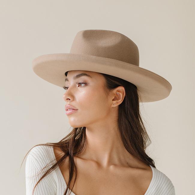 Woman wearing a tan pencil brim fedora hat and a white top, facing sideways against a plain background #color_tan