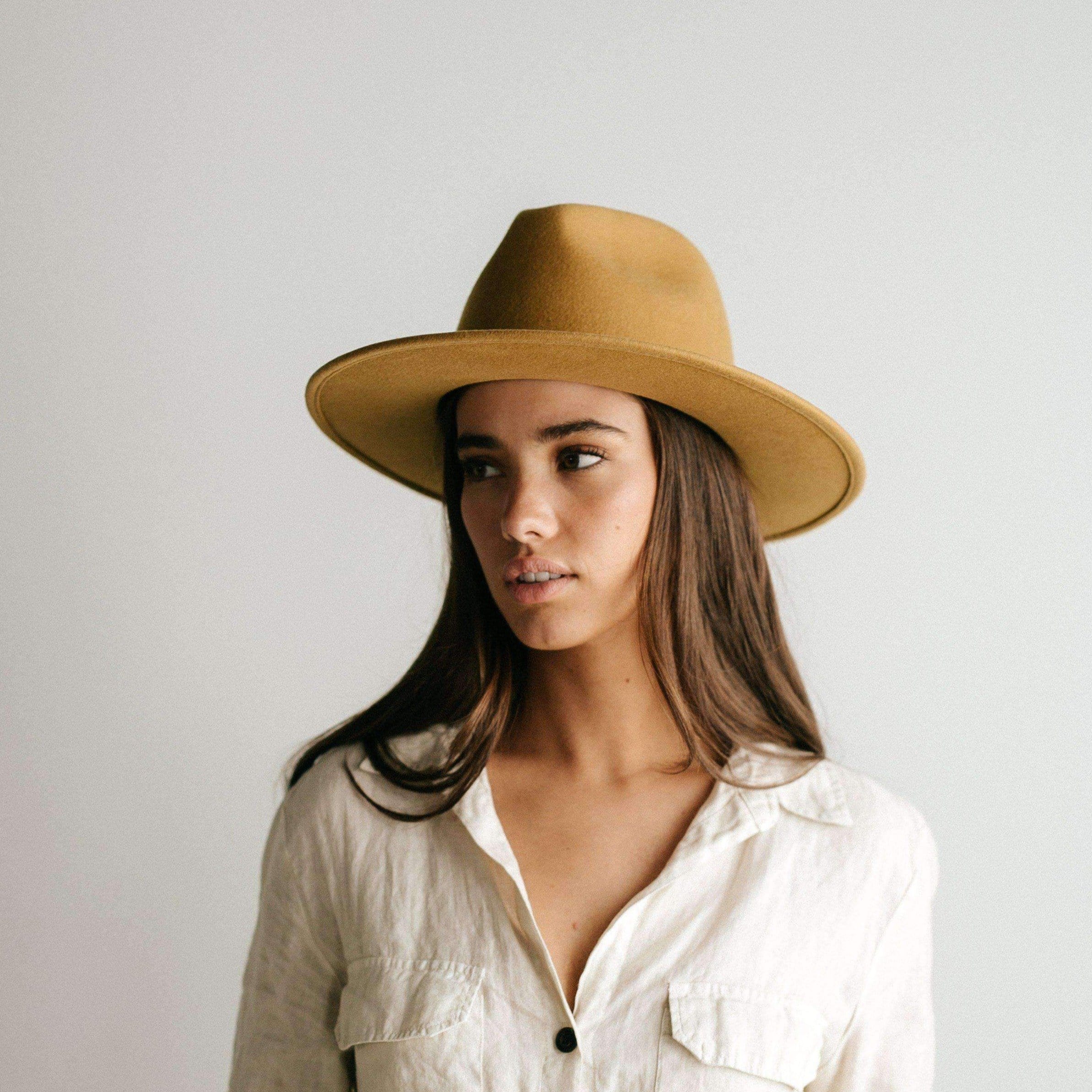 Woman wearing a mustard colored fedora hat and a white shirt, looking sideways against a plain background #color_mustard