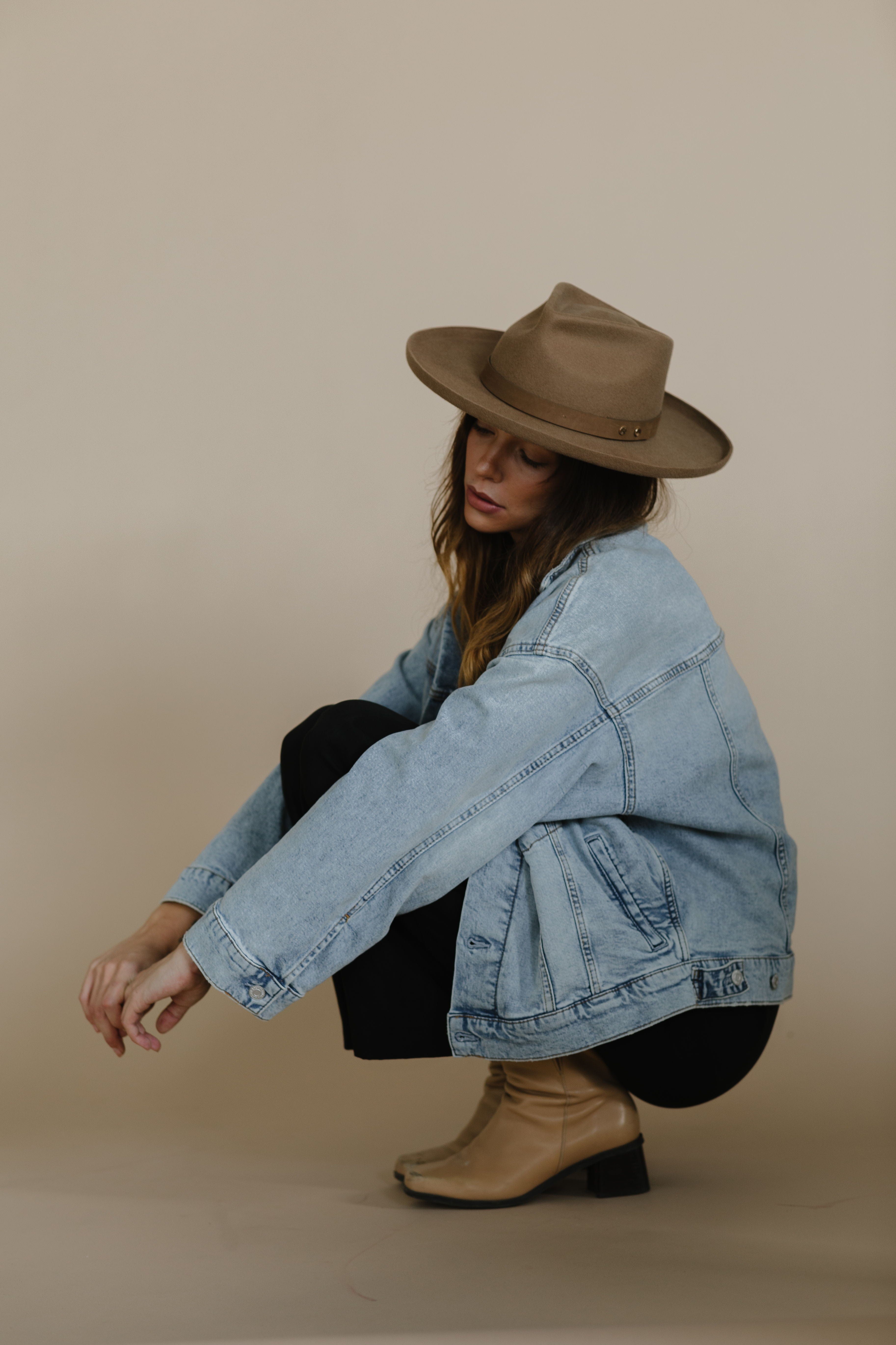 Woman wearing a tan pencil brim fedora hat with a hat band, a denim jacket, black pants and tan boots, sitting against a plain background #color_tan